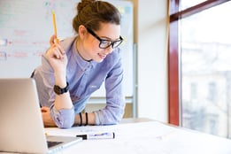 Happy young woman in glasses standing near the window in office and working with blueprint.jpeg
